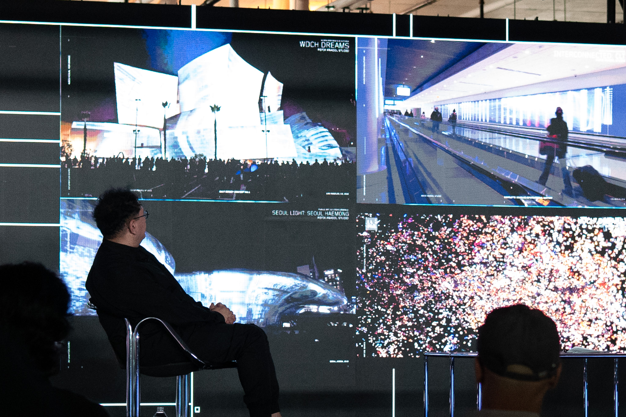 Refik Anadol seated on stage in silhouette before a vast wall showing his works WDCH Dreams and Seoul Light