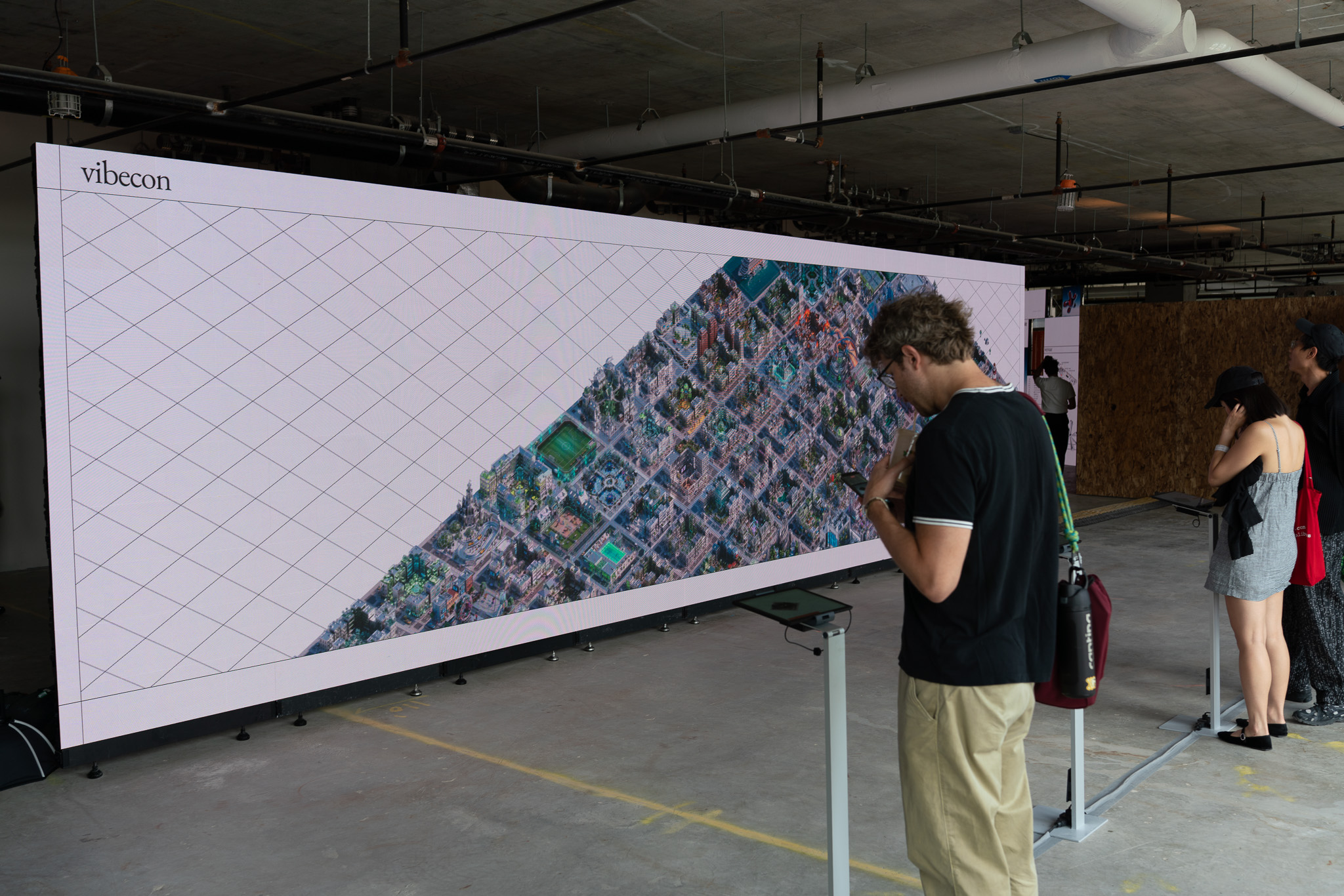 A long vibecon LED wall showing a dense pixel-art city, with attendees adding to it from tablets