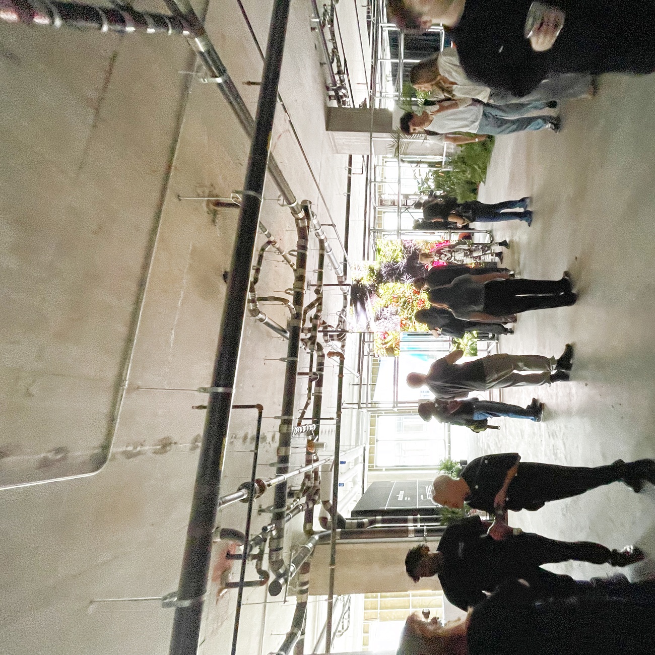 Attendees moving through the raw concrete floor beneath exposed pipes, a lush planted installation glowing at the back