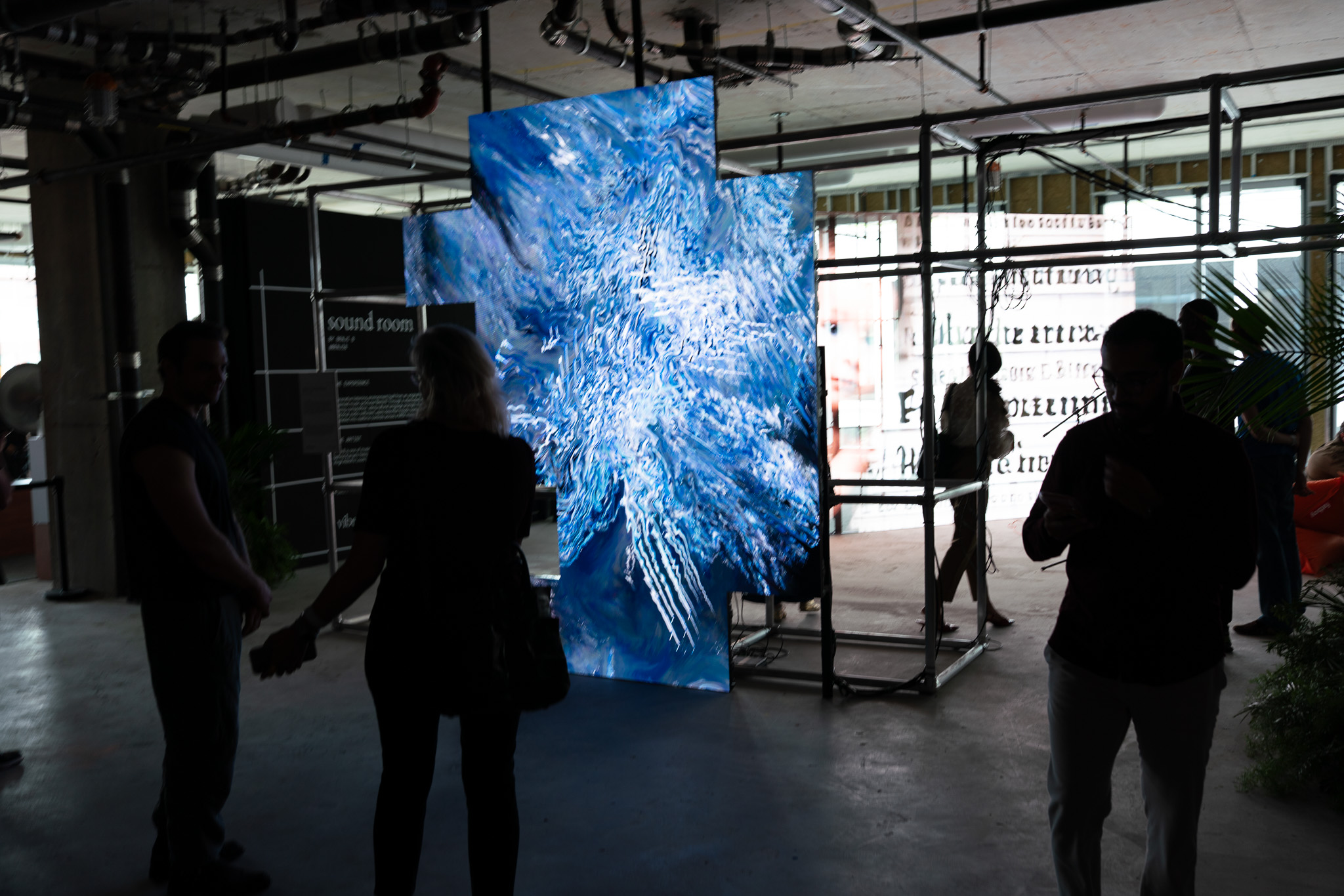 A tall screen of swirling blue brushwork glowing in the dark raw space, visitors in silhouette before it