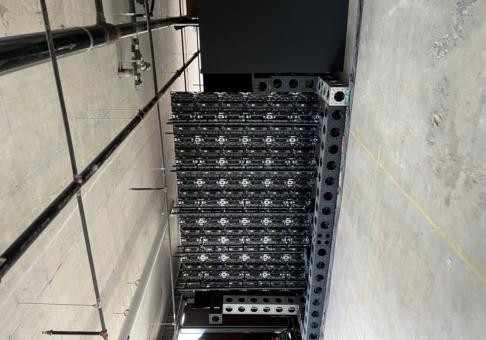 The back of a massive LED video wall and its scaffolding standing in a raw concrete floor with exposed ceiling pipes