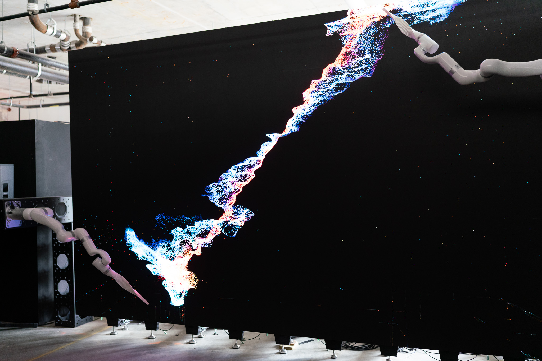 Two robotic arms drawing a luminous particle trail diagonally across a vast black LED wall