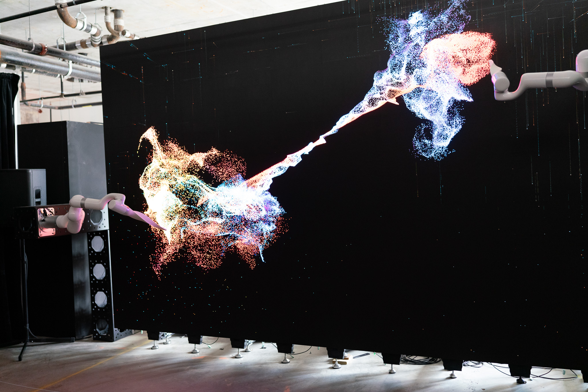 Robotic arms flinging bursts of colored particles across the black wall