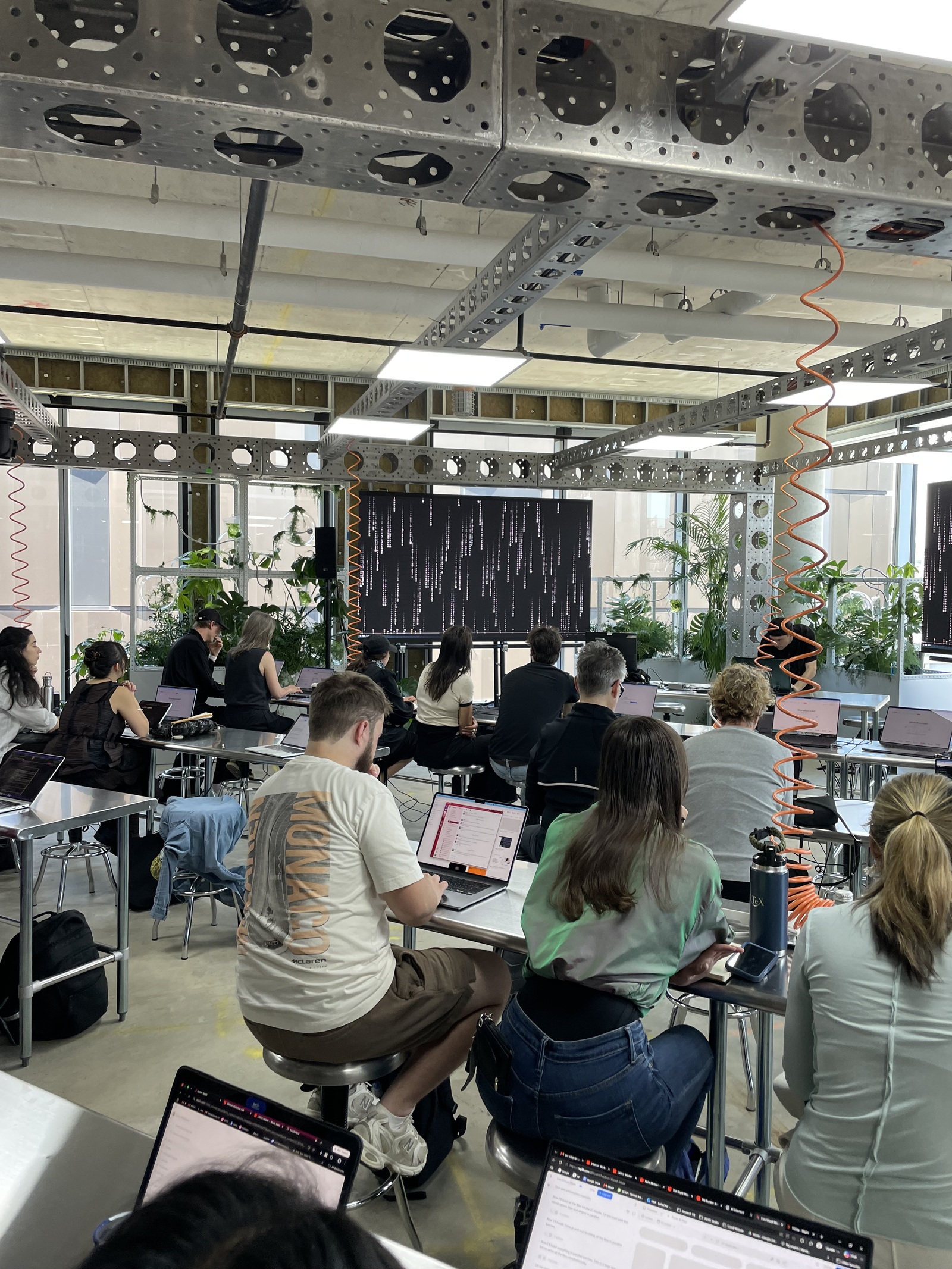 People at steel workbenches on laptops in the lab room, a big screen showing Matrix-style code rain, daylight and plants behind