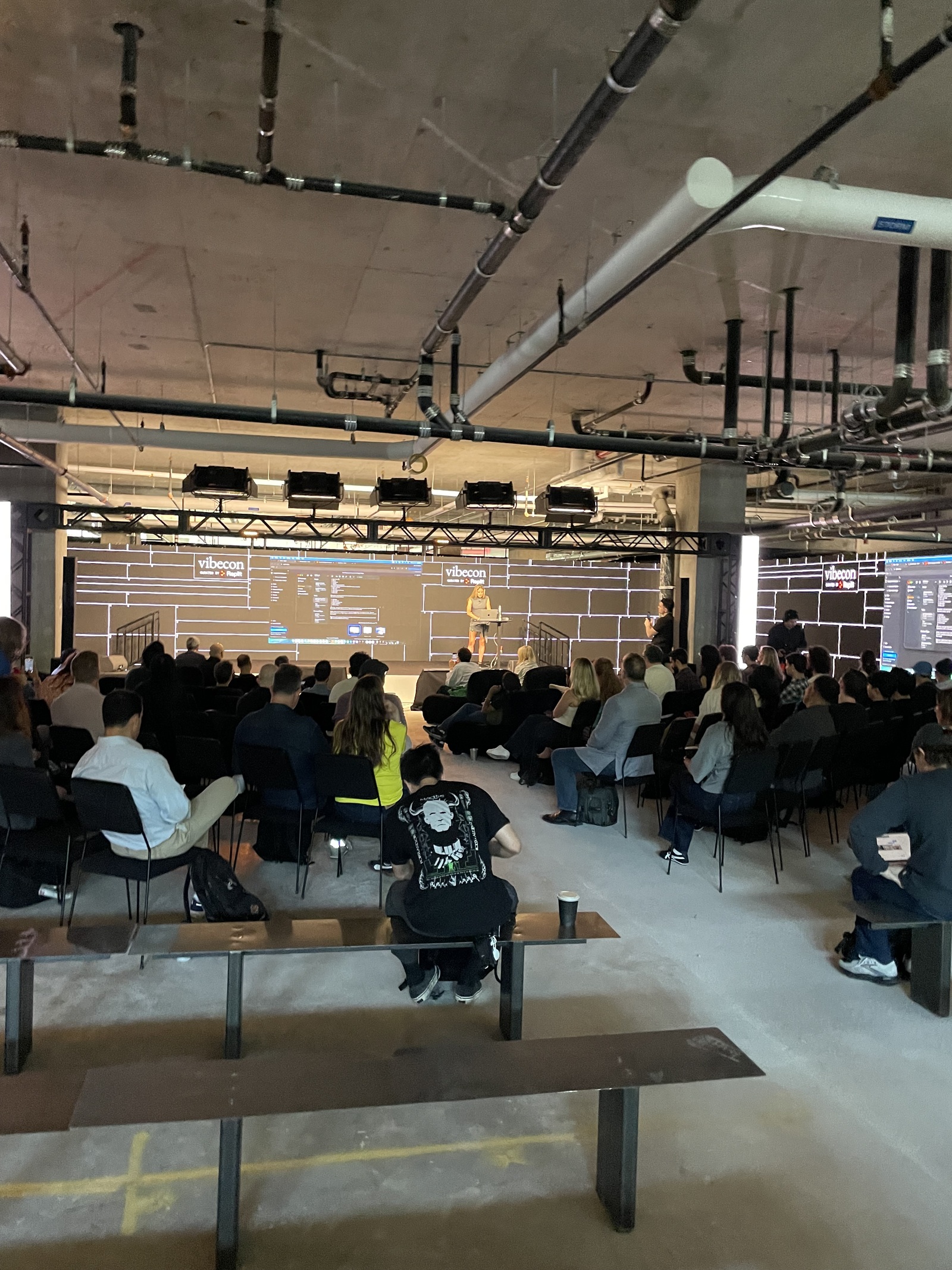 A speaker at a laptop on the vibecon LED-wall stage, the audience seated in rows on the raw concrete floor beneath exposed pipes