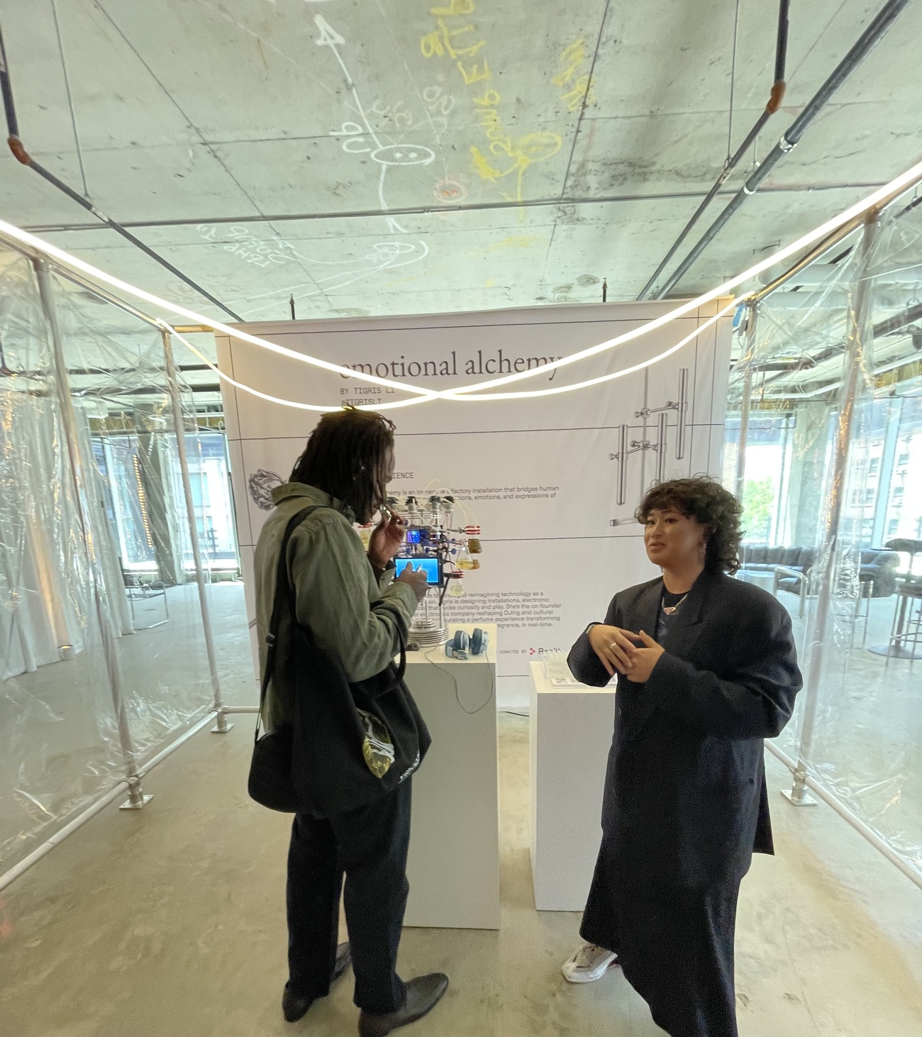 Tigris Li speaking with a visitor at the Emotional Alchemy booth in the raw, plastic-draped space